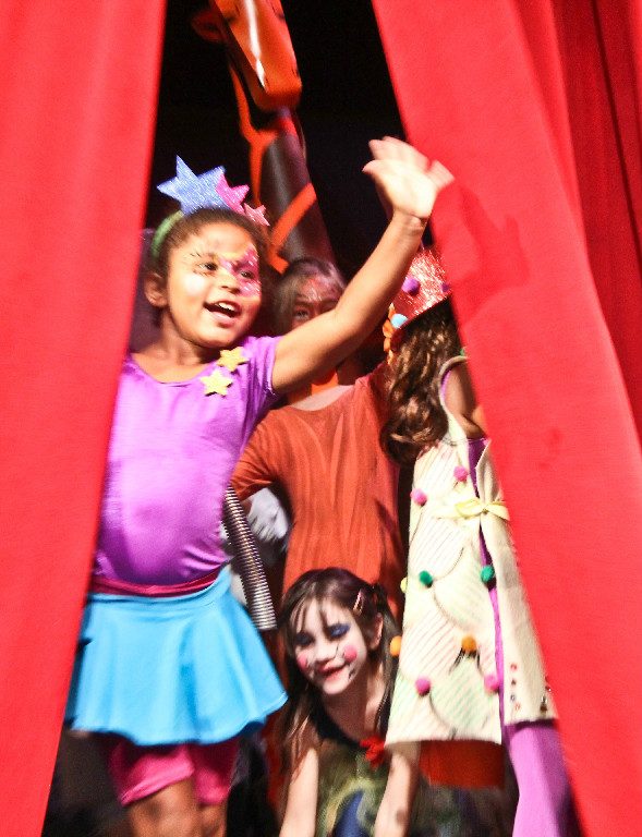 A young performer waves from between red stage curtains